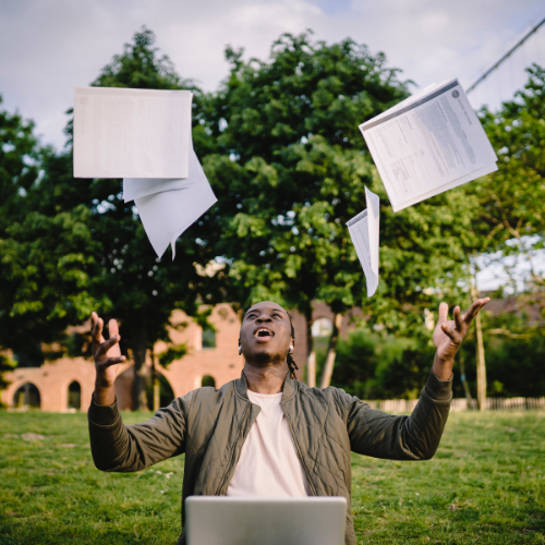 Man standing in front of a laptop outside throwing paper in the air.