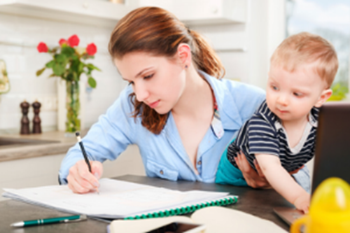 Young woman holding a toddler while making notes in at a kitchen table