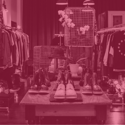 A clothes shop with a display of shoes on a table and clothes hanging on racks in the background. Image has a red tint.