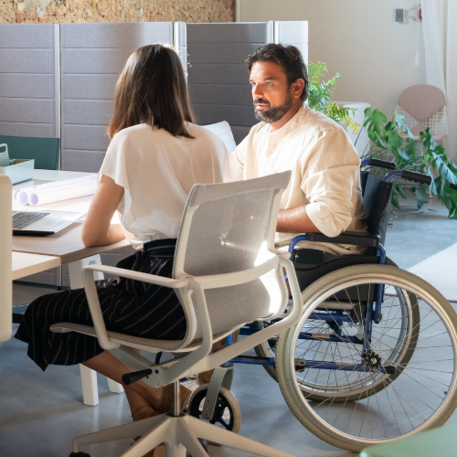 Two people sitting at a desk. One woman sitting on a chair and one man in a wheelchair.