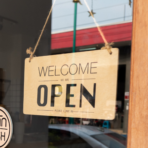 A wooden sign hanging in a door window. The sign says welcome, we are open, please come in.